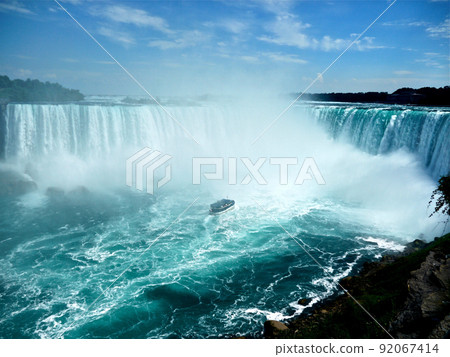 Niagara Falls. A cruise ship surrounded by fog in a basin. A large amount of water falling from a waterfall that spreads across the width. 92067414