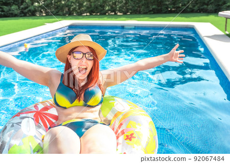 young red-haired woman with hat and sunglasses, shouting with joy, arms raised, sitting on a float in the pool. happy young woman on summer holiday. concept of holidays and travel. young red-haired woman with hat and sunglasses, shouting with joy, arms raised, sitting on a float in the pool. happy young woman on summer holiday. concept of holidays and travel. 92067484