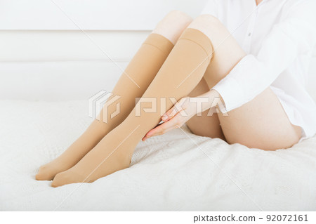 Beige compression stockings on a woman in a white room. Knee socks or socks. Girl putting on stockings at home. Beautiful female legs. Beige compression stockings on a woman in a white room. Knee socks or socks. Girl putting on stockings at home. Beautiful female legs. 92072161