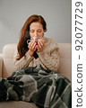 A woman sitting on the couch drinks hot tea for illness. 92077729
