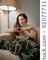 Woman sits on a sofa with a mug of healing tea on a winter evening. 92077731