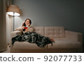 Woman with a cup of cough tea sits in a blanket on the sofa at home. 92077733