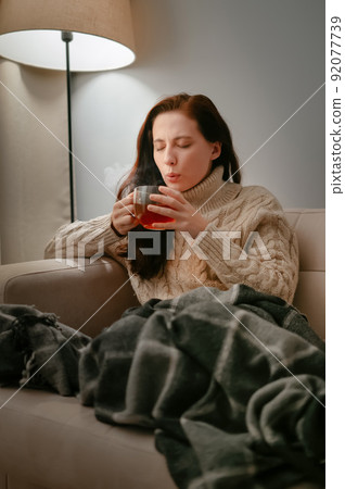 Woman at home is treated with hot tea while sitting on a beige sofa. Woman at home is treated with hot tea while sitting on a beige sofa. 92077739