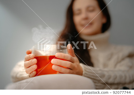Woman warms her hands on a glass mug with tea on winter evening home. Woman warms her hands on a glass mug with tea on winter evening home. 92077744