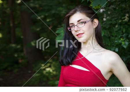 young brunette woman in a red dress and glasses in city park 92077982