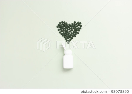 Open vitamin jar with easy to swallow pills of natural green spirulina in heart shape, light green background, with copy space. They are made of blue-green algae with strong antioxidant effects. Open vitamin jar with easy to swallow pills of natural green spirulina in heart shape, light green background, with copy space. They are made of blue-green algae with strong antioxidant effects. 92078890