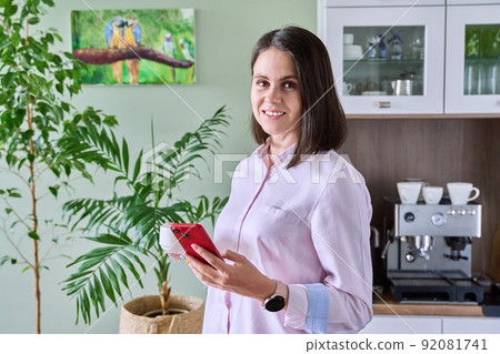 Young woman in kitchen with cup of coffee smartphone, looking at camera Young woman in kitchen with cup of coffee smartphone, looking at camera 92081741