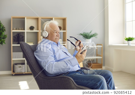 Senior man sitting in armchair at home wearing headphones and using mobile phone 92085412