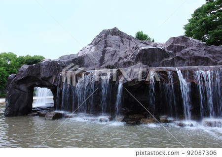 A park with a waterfall. The sound of the water falling from the rocks is intense, and the water play area in the park is waiting for children. A park with a waterfall. The sound of the water falling from the rocks is intense, and the water play area in the park is waiting for children. 92087306