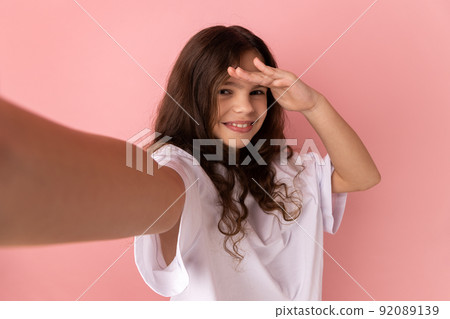 Portrait of smiling satisfied little girl wearing white T-shirt looking into distance with positive facial expression, point of view. Indoor studio shot isolated on pink background. 92089139