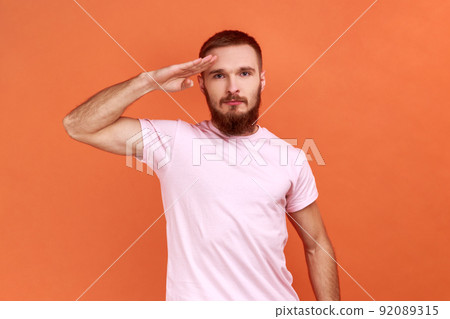 Yes sir. Portrait of serious strict bearded man standing as soldier giving salute, ready for order, wearing pink T-shirt. Indoor studio shot isolated on orange background. 92089315
