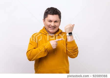 Good result and on time. Positive man standing, showing his smart watch and looking at camera with toothy smile, wearing urban style hoodie. Indoor studio shot isolated on white background. Good result and on time. Positive man standing, showing his smart watch and looking at camera with toothy smile, wearing urban style hoodie. Indoor studio shot isolated on white background. 92089366