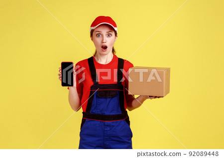 Shocked astonished delivery woman standing with package and showing cell phone with empty display, keeps mouth open, wearing overalls and red cap. Indoor studio shot isolated on yellow background. 92089448