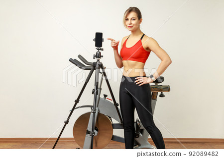 Beautiful slim woman standing near exercise bike, pointing at phone on tripod and looking smiling at camera, wearing sports tights and red top. Indoor studio shot on gray wall background. Beautiful slim woman standing near exercise bike, pointing at phone on tripod and looking smiling at camera, wearing sports tights and red top. Indoor studio shot on gray wall background. 92089482