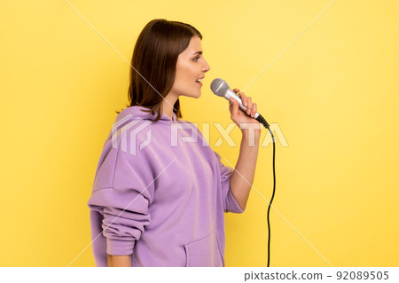 Side view of young beautiful woman reporter holding microphone, reporting and telling opinion, discussing important topics, wearing purple hoodie. Indoor studio shot isolated on yellow background. 92089505