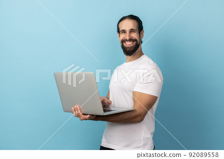 Portrait of man wearing white T-shirt standing with portable computer in hand, looking at camera with toothy smile, watching movie or video. Indoor studio shot isolated on blue background. Portrait of man wearing white T-shirt standing with portable computer in hand, looking at camera with toothy smile, watching movie or video. Indoor studio shot isolated on blue background. 92089558