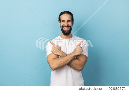 Portrait of positive satisfied man wearing white T-shirt standing with crossed hands and pointing to copy space for advertisement on both sides. Indoor studio shot isolated on blue background. 92089571