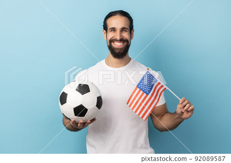 Portrait of satisfied delighted man wearing white T-shirt holding flag of united states of america and soccer black and white ball, united soccer league. Indoor studio shot isolated on blue background Portrait of satisfied delighted man wearing white T-shirt holding flag of united states of america and soccer black and white ball, united soccer league. Indoor studio shot isolated on blue background 92089587