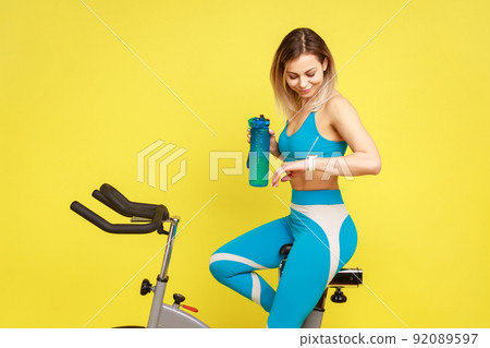 Sporty woman riding on exercise bike with water bottle in hands and looking at her smartwatch, checking heart beat, wearing blue sportswear. Indoor studio shot isolated on yellow background. Sporty woman riding on exercise bike with water bottle in hands and looking at her smartwatch, checking heart beat, wearing blue sportswear. Indoor studio shot isolated on yellow background. 92089597