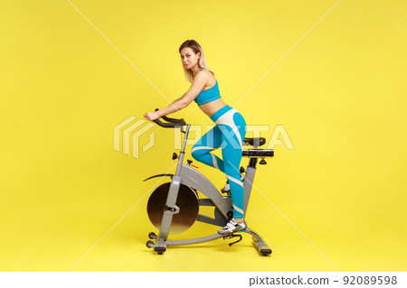 Side view full length of blonde beautiful woman riding exercise bike, healthy lifestyle and motivation, wearing blue sportswear. Indoor studio shot isolated on yellow background. Side view full length of blonde beautiful woman riding exercise bike, healthy lifestyle and motivation, wearing blue sportswear. Indoor studio shot isolated on yellow background. 92089598