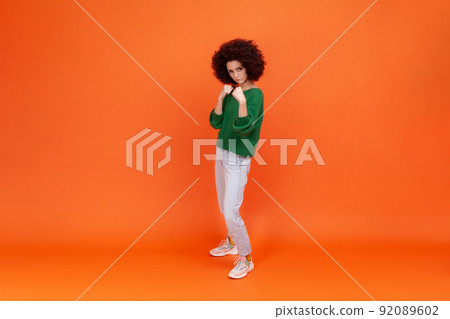 Full length portrait of woman with Afro hairstyle wearing green casual style sweater and jeans, standing with raised fists, expressing anger. Indoor studio shot isolated on orange background. 92089602