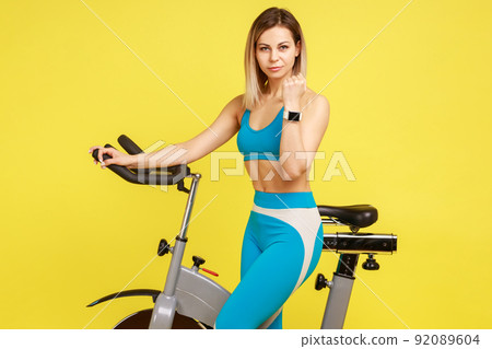 Portrait of blonde woman doing cardio exercises, standing and showing her smartwatch or fitness tracker, wearing blue sportswear. Indoor studio shot isolated on yellow background. 92089604