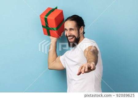 Portrait of happy satisfied positive bearded man wearing white T-shirt being ready to throw birthday box, having fun and sharing gift. Indoor studio shot isolated on blue background. 92089625
