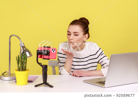 Romantic woman blogger in striped shirt recording video for followers, air sending kissing over palms hands, expressing love. Indoor studio studio shot isolated on yellow background Romantic woman blogger in striped shirt recording video for followers, air sending kissing over palms hands, expressing love. Indoor studio studio shot isolated on yellow background 92089677