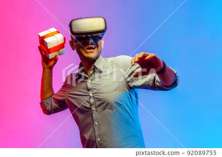 Portrait of smiling happy man in shirt wearing vr headset, throwing present box, having excited facial expression. Indoor studio shot isolated on colorful neon light background. 92089755