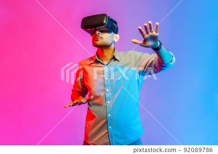 Portrait of concentrated man in shirt discovering new technologies wearing virtual reality headset, futuristic 3d vision. Indoor studio shot isolated on colorful neon light background. 92089786