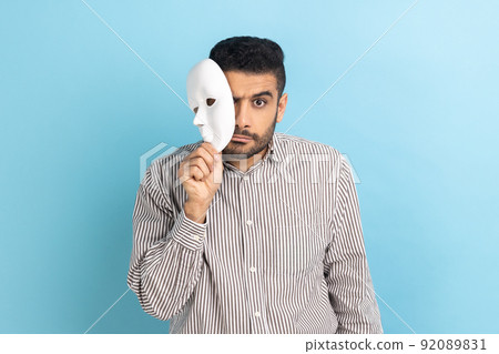 Serious bearded businessman removing white mask from face showing his strict expression, pretending to be another person, wearing striped shirt. Indoor studio shot isolated on blue background. 92089831