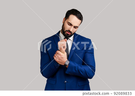Portrait of businessman standing with grimace of pain, massaging sore wrist, suffering hand injury or sprain, wearing official style suit. Indoor studio shot isolated on gray background. Portrait of businessman standing with grimace of pain, massaging sore wrist, suffering hand injury or sprain, wearing official style suit. Indoor studio shot isolated on gray background. 92089833