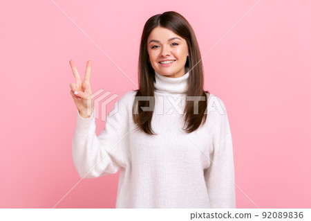 Positive dark haired young female smiling happily and showing v sign or peace gesture, rejoicing win, wearing white casual style sweater. Indoor studio shot isolated on pink background. Positive dark haired young female smiling happily and showing v sign or peace gesture, rejoicing win, wearing white casual style sweater. Indoor studio shot isolated on pink background. 92089836