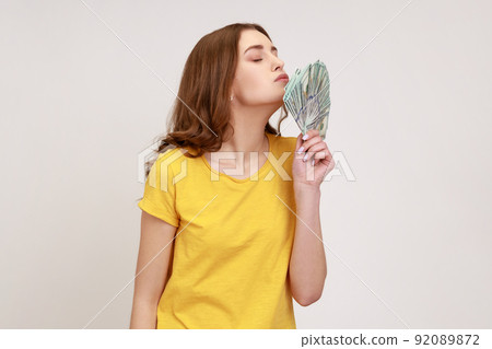 Side view portrait of cheerful rich attractive young woman in yellow casual style T-shirt standing and air kissing fan of dollars, happy to win lottery. Indoor studio shot isolated on gray background. 92089872