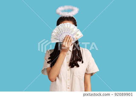 Angelic rich woman with black dreadlocks and nimbus on head hiding behind dollar banknotes, big profit earning cash, wearing white shirt. Indoor studio shot isolated on blue background. 92089901