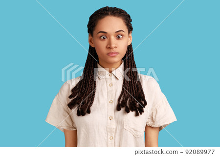 Portrait of funny silly woman with black dreadlocks looking at camera, cross eyed with stupid dumb face, has awkward confused comical expression. Indoor studio shot isolated on blue background. 92089977