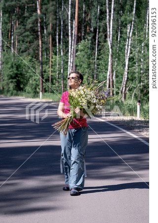 full-length portrait of a happy smiling woman with a huge bouquet of wild flowers walking along a forest road 92096153