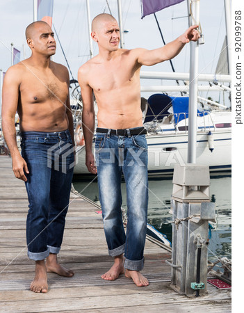 two guys without shirts in jeans walk barefoot by the handle near yachts in the port two guys without shirts in jeans walk barefoot by the handle near yachts in the port 92099788