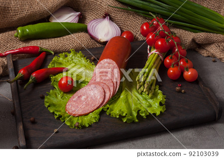 smoked servelat on a wooden board decorated with fresh vegetables smoked servelat on a wooden board decorated with fresh vegetables 92103039