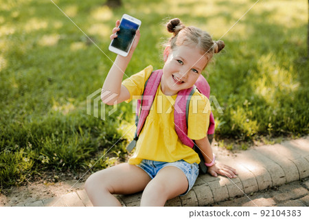 A charming little girl with a backpack is sitting on the sidewalk after school and showing her phone. Back to school, after school, school age A charming little girl with a backpack is sitting on the sidewalk after school and showing her phone. Back to school, after school, school age 92104383