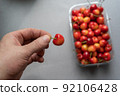A man's hand holds a ripe sweet cherry berry.  A new crop of red berries in a transparent tray in the background. Healthy Food. Selective focus. 92106428