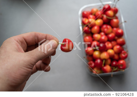 A man's hand holds a ripe sweet cherry berry. A new crop of red berries in a transparent tray in the background. Healthy Food. Selective focus. A man's hand holds a ripe sweet cherry berry. A new crop of red berries in a transparent tray in the background. Healthy Food. Selective focus. 92106428