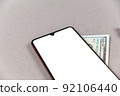 Smartphone and money on a gray screen. Smartphone with a white blank screen. A hundred-dollar bill lies under the phone. Copy space for text and design elements. Selective focus. 92106440