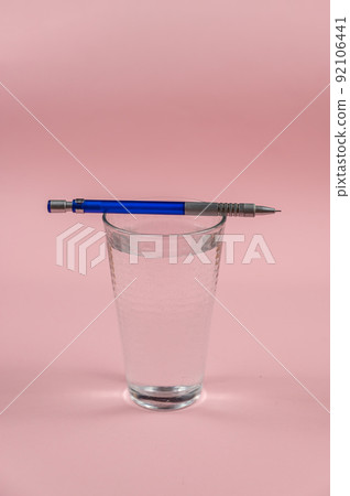 A glass of water and a pencil on a pink background. A full glass of drinking water. Blue and silver metallic pencil. Kurp plan. Selective focus. A glass of water and a pencil on a pink background. A full glass of drinking water. Blue and silver metallic pencil. Kurp plan. Selective focus. 92106441
