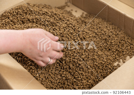 A woman hand picks up dry cat food. The food is in bulk in an open cardboard box. Triangular brown pellets. Pet supplies. Selective Focus. 92106443