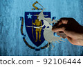 Witch flying on broomstick in front of Commonwealth of Massachusetts flag. Man hand holding figurine cut out of paper. Celebrating All Saints Night in United States concept. Selective focus. 92106444