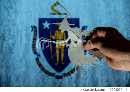 Witch flying on broomstick in front of Commonwealth of Massachusetts flag. Man hand holding figurine cut out of paper. Celebrating All Saints Night in United States concept. Selective focus. Witch flying on broomstick in front of Commonwealth of Massachusetts flag. Man hand holding figurine cut out of paper. Celebrating All Saints Night in United States concept. Selective focus. 92106444