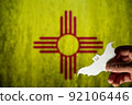 Happy Halloween, New Mexico. Male hand holds bat figurine. New Mexico state flag in background. Cut out of white paper Halloween paraphernalia. All Hallows evening. Selective focus. 92106446