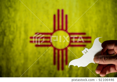 Happy Halloween, New Mexico. Male hand holds bat figurine. New Mexico state flag in background. Cut out of white paper Halloween paraphernalia. All Hallows evening. Selective focus. Happy Halloween, New Mexico. Male hand holds bat figurine. New Mexico state flag in background. Cut out of white paper Halloween paraphernalia. All Hallows evening. Selective focus. 92106446
