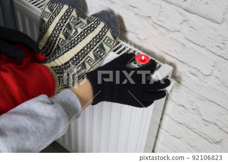 A woman's hand in winter gloves warms her hands from a heating battery. The concept of the cost of heating services, the increase in prices for utilities. Lack of heat. A woman's hand in winter gloves warms her hands from a heating battery. The concept of the cost of heating services, the increase in prices for utilities. Lack of heat. 92106823